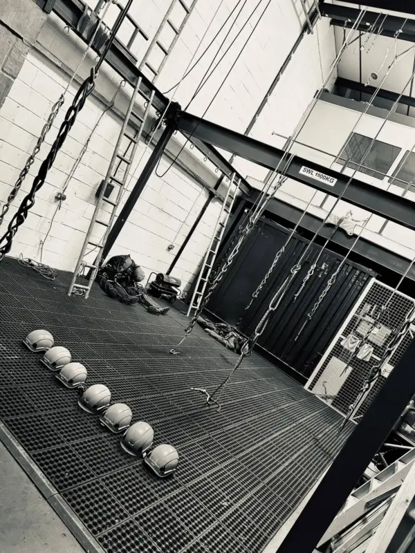 A black and white photo of the interior of a rope access training facility, showing a large steel training rig with ladders and ropes hanging from the ceiling. A row of hard hats is lined up on the grated metal floor.