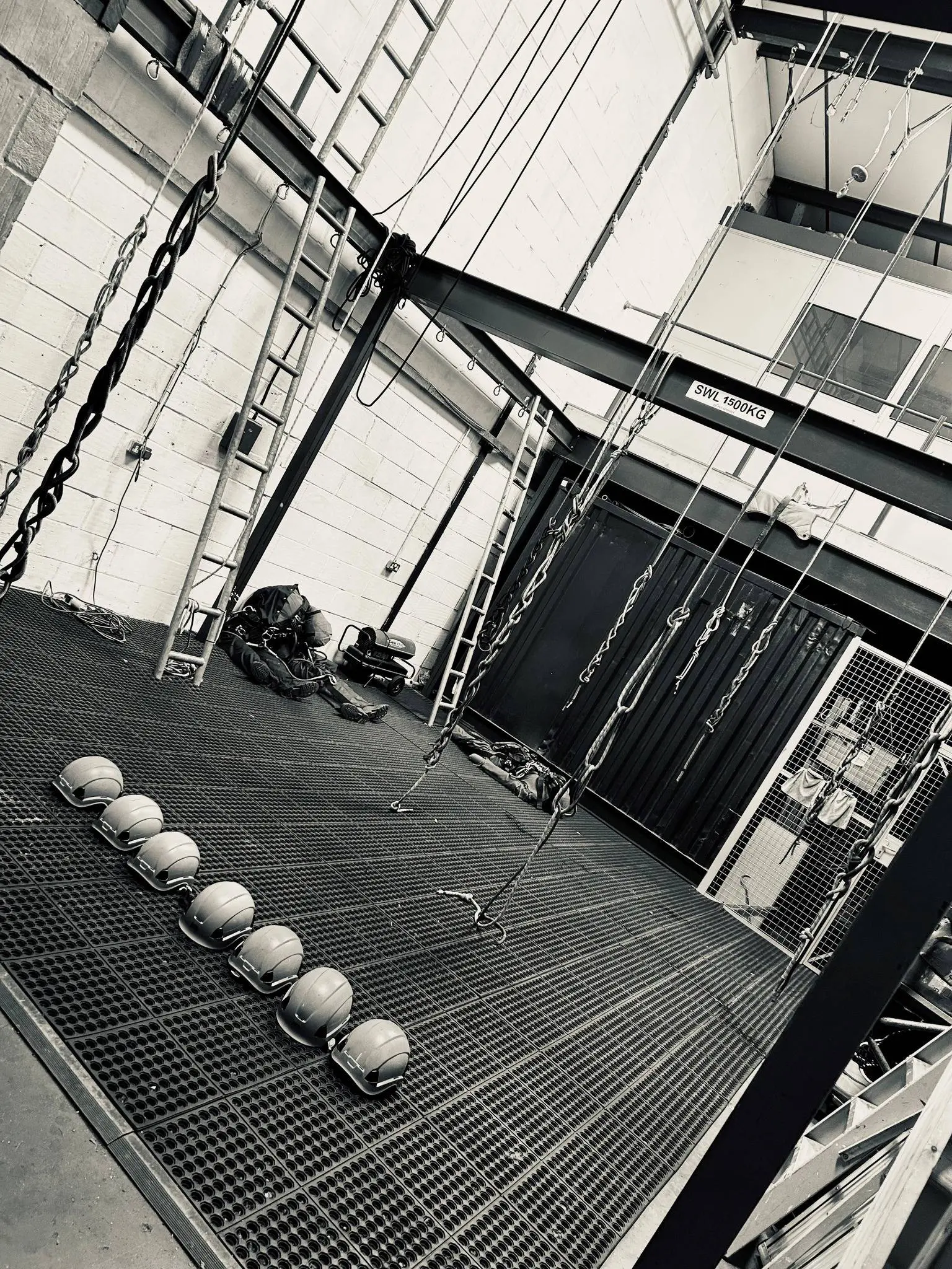 A black and white photo of the interior of a rope access training facility, showing a large steel training rig with ladders and ropes hanging from the ceiling. A row of hard hats is lined up on the grated metal floor.