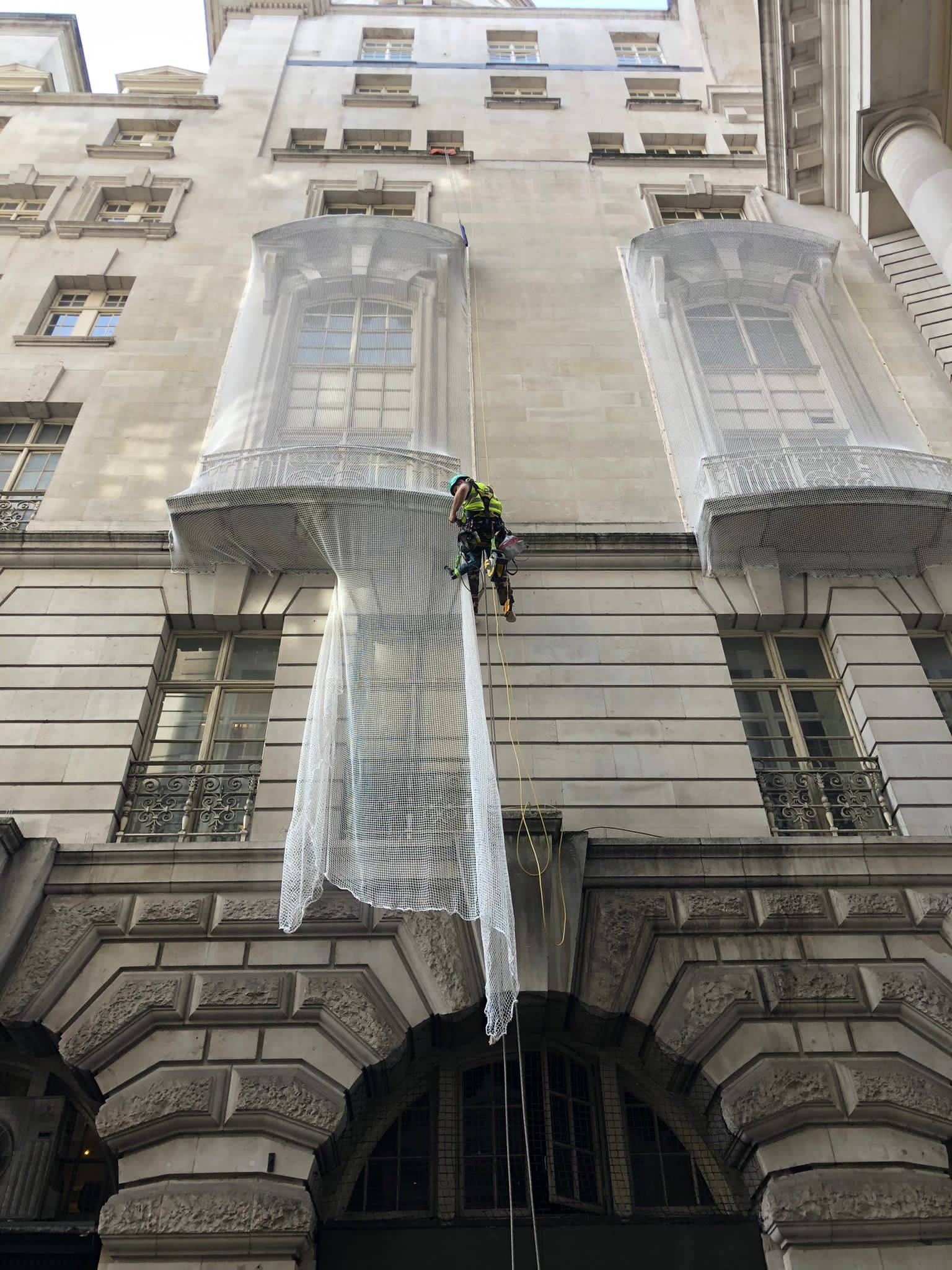 AAA rope technician in safety gear installing safety nets on a damaged building facade, performing stone repairs and addressing spalling concrete.
