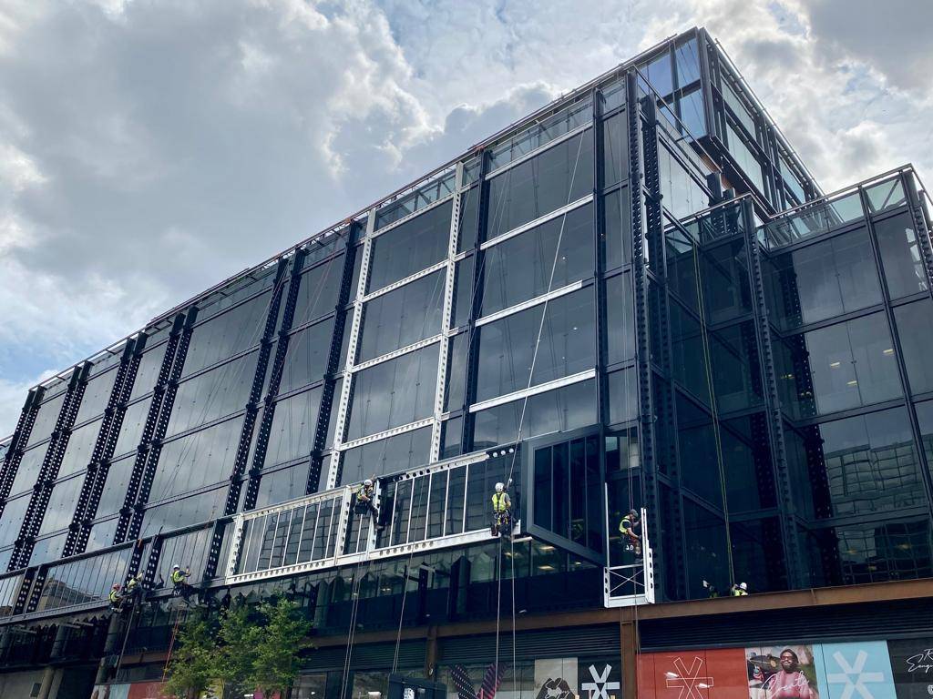AAA rope technician in safety gear painting the superstructure of Buckingham Palace Road, changing from white to anthracite with London buildings in the background.