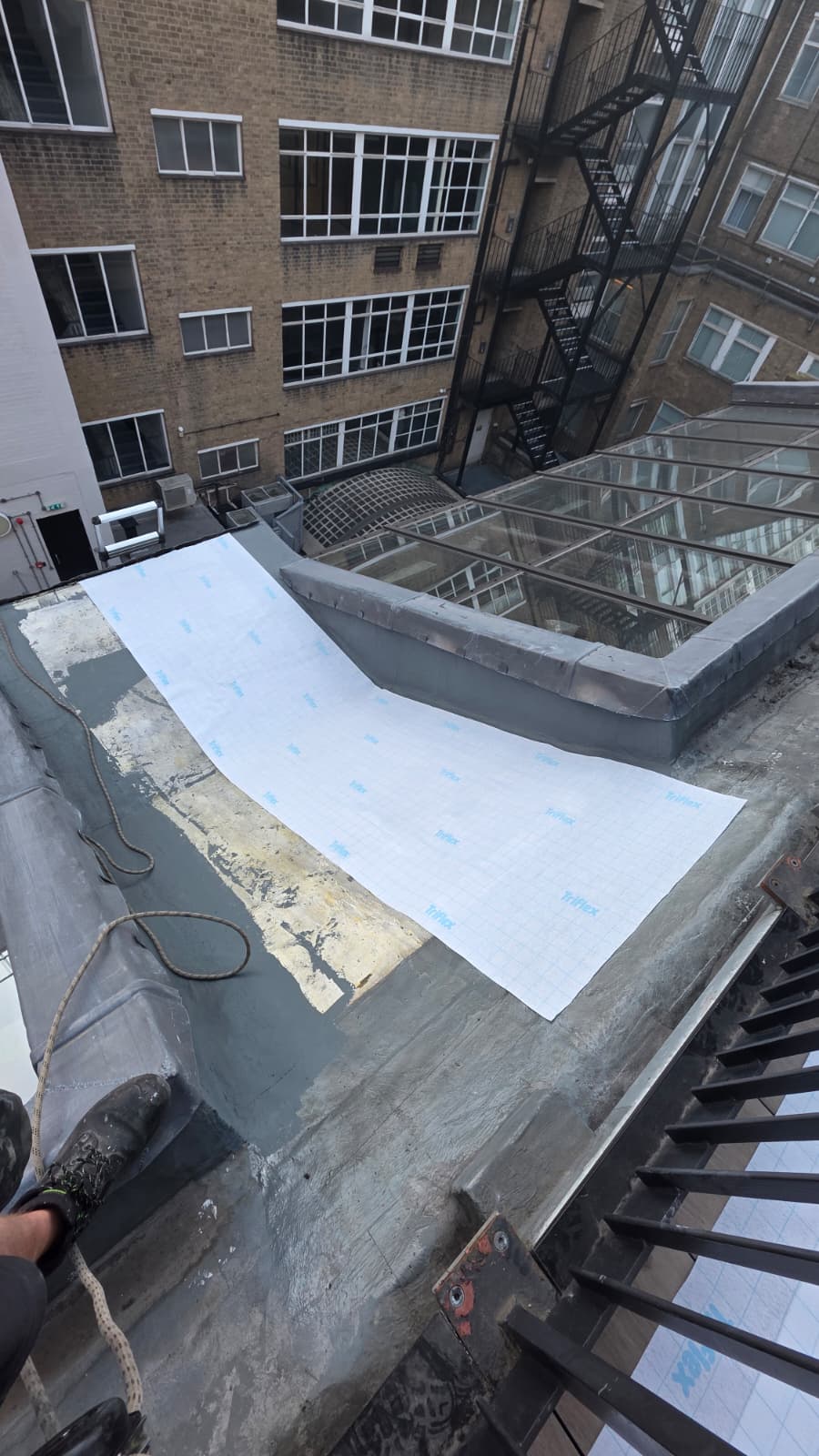 AAA rope technician in safety gear applying Tri-Flex fleece on a leaking sloped roof in a well area, with dormer roof visible.