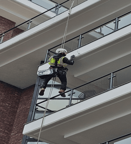 AAA rope technician in safety gear painting and repairing masonry, balconies, and brickwork at Waterside Point with a London river view in the background.